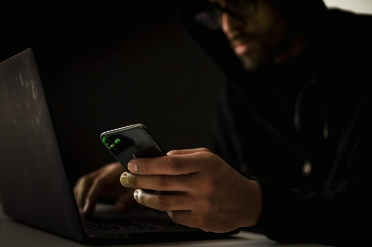 crop ethnic hacker with smartphone typing on laptop in dark room